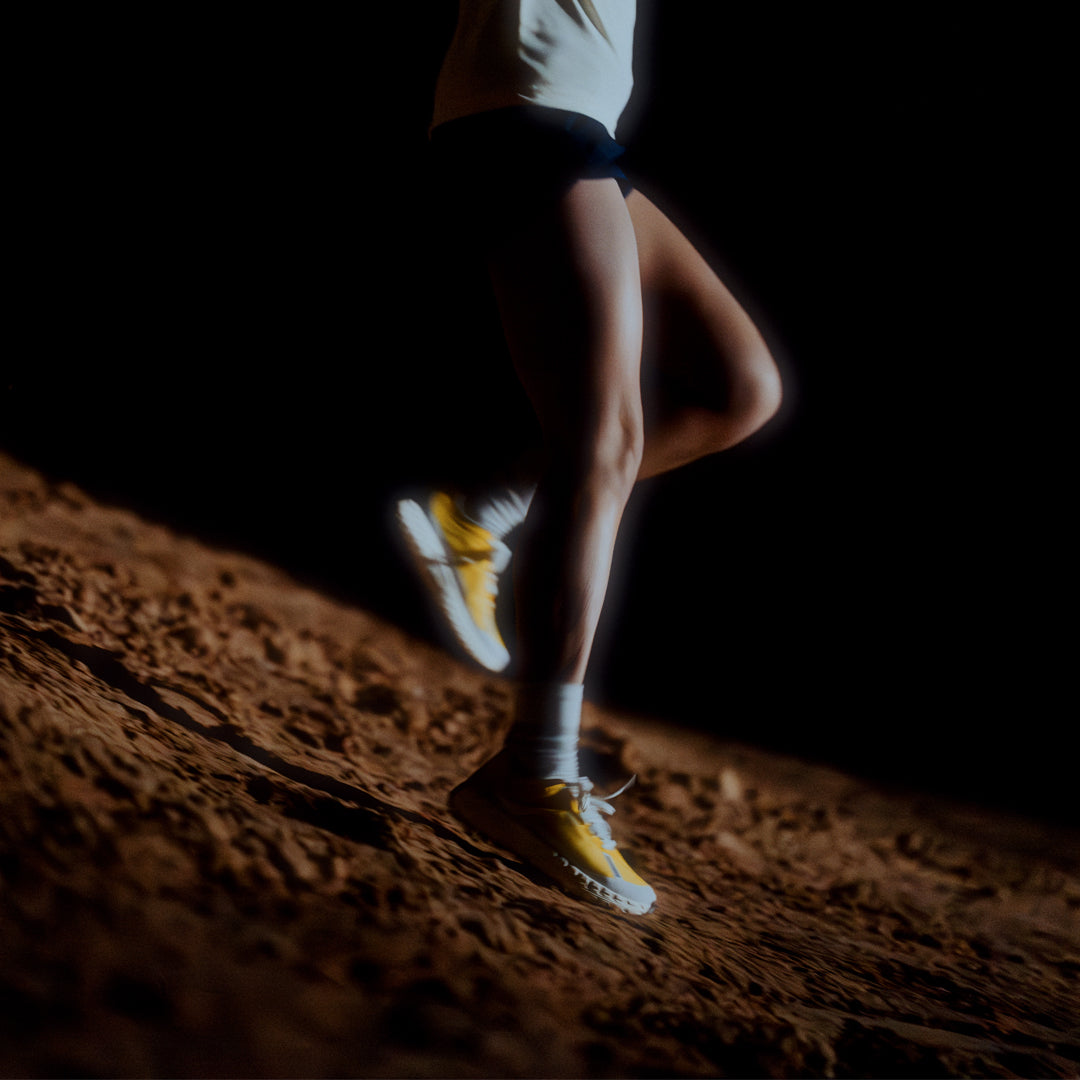 norda-001-trail-shoes-parhelion-womens-thepace-lifestyle-shot of a person's legs running against a dark background and rocky terrain wearing the norda shoes