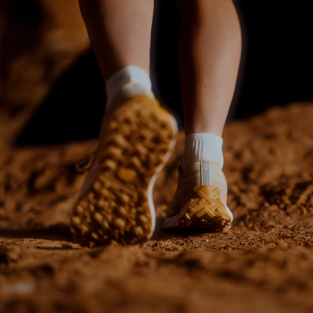 norda-002-trail-running-shoe-men's-Sand-available-at-thepace-lifestyle-shot of the back of the shoes with soles visible worn by a person walking away form teh camera on clay terrain with white socks